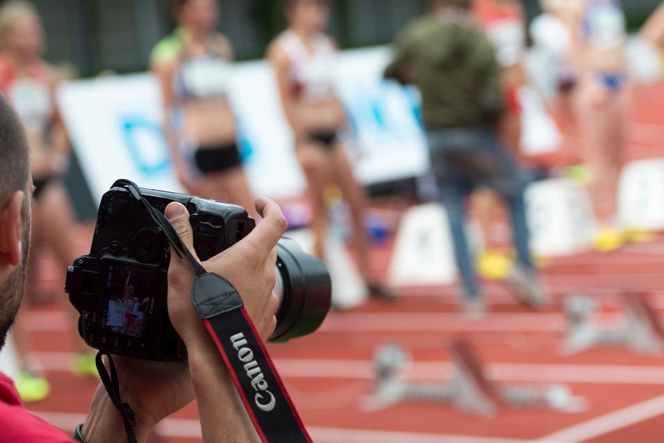 Fotograf mit Kamera im Vordergrund, im Hintergrund stehen Athletinnen an einer Laufbahn.