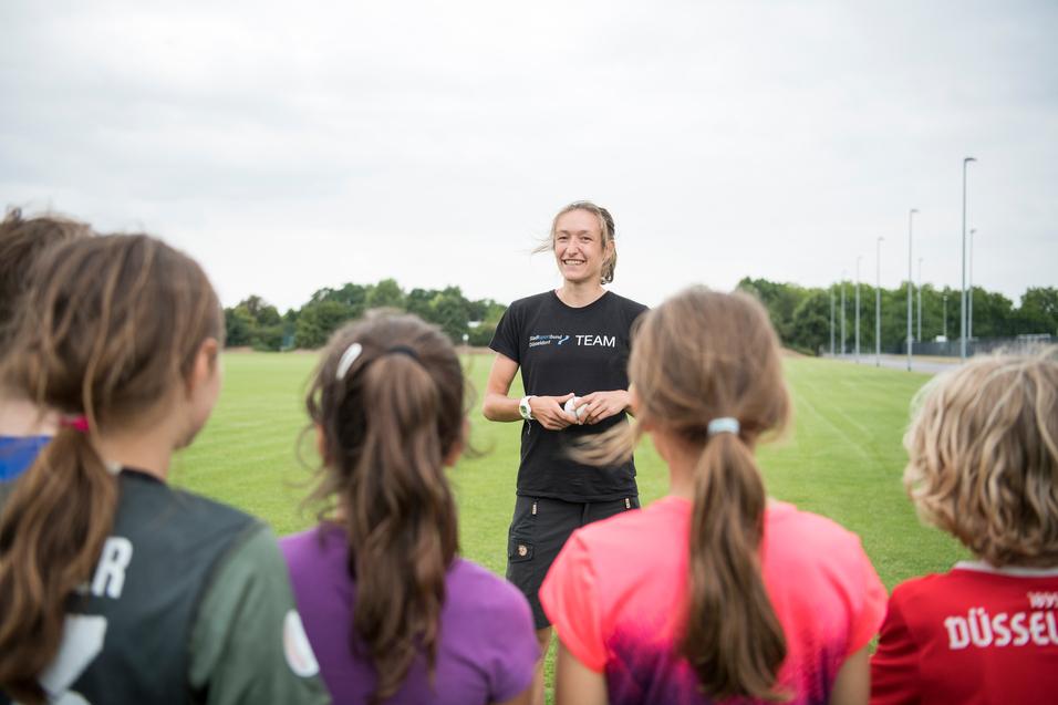 Trainerin spricht mit jungen Sportlerinnen auf einem grünen Fußballfeld unter bewölkten Himmel.