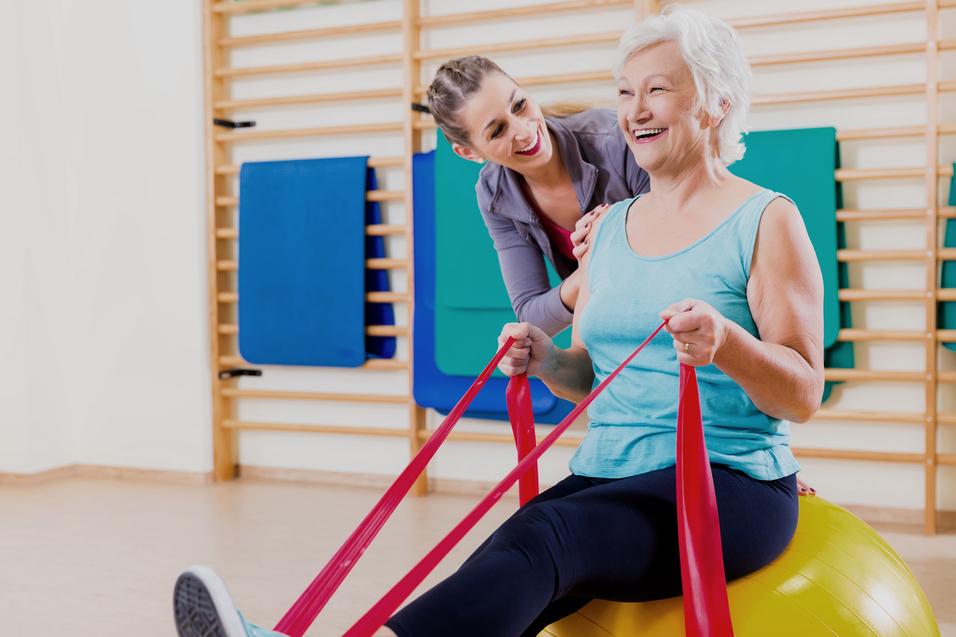 Frau mit grauen Haaren sitzt auf einem großen Gymnastikball, während eine Trainerin ihr mit engen Bändern hilft.