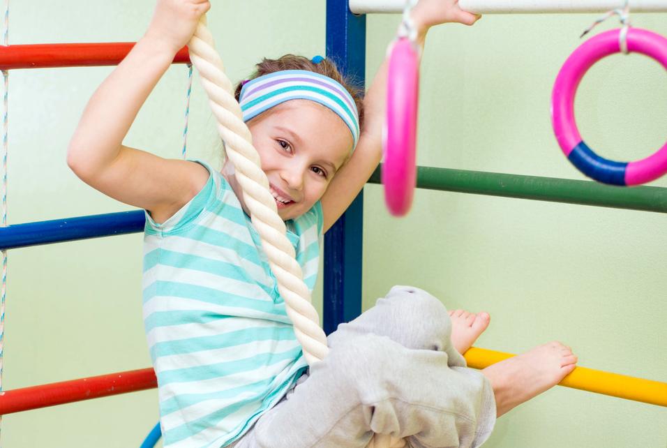 Ein Mädchen hängt lachend an einem Kletterseil in einer Turnhalle, umgeben von bunten Turngeräten.